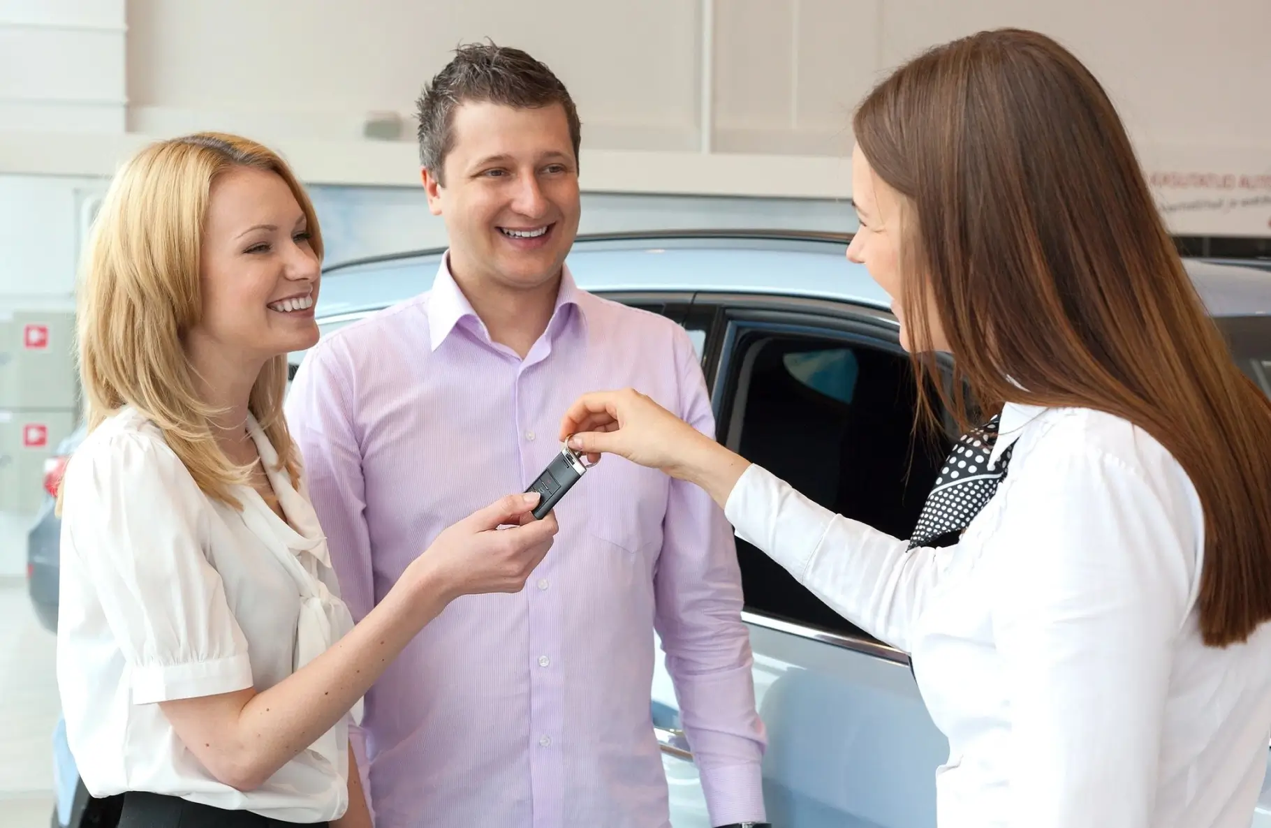 A woman handing keys to two people in front of a car.